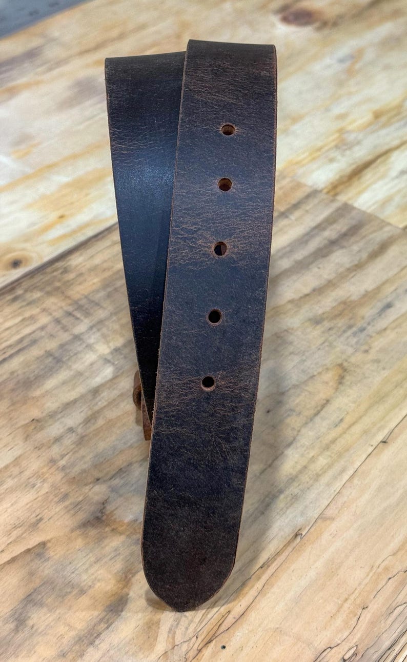 May include: A dark brown leather belt with a distressed, textured finish. The belt features a rounded tip and a series of five evenly spaced circular holes punched along the center for adjustment, showcasing the natural grain of the material.