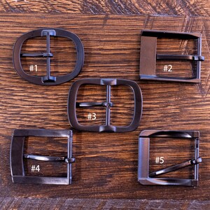May include: Five dark metal belt buckles in various shapes: oval, rectangular, and square. Each buckle has a bar for the belt strap and is numbered from one to five. The buckles are displayed on a wooden surface.