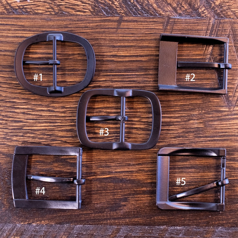 May include: Five dark metal belt buckles in various shapes: oval, rectangular, and square. Each buckle has a bar for the belt strap and is numbered from one to five. The buckles are displayed on a wooden surface.