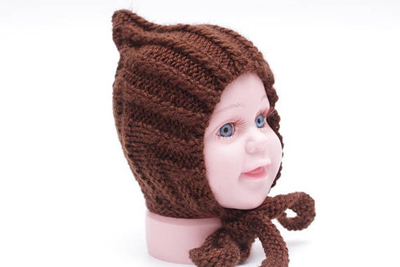 K&ouml;nnte beinhalten: Eine braune gestrickte Baby-M&uuml;tze mit einer spitzen Spitze und einer langen Schleife. Die M&uuml;tze ist aus weichem, dickem Garn gefertigt und hat ein schlichtes, klassisches Design.