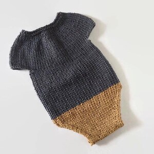 May include: A handmade, two-tone knitted baby romper. The top portion is a dark gray, and the bottom is a warm tan color. The romper has short sleeves and a round neckline. The knit fabric has a textured appearance.