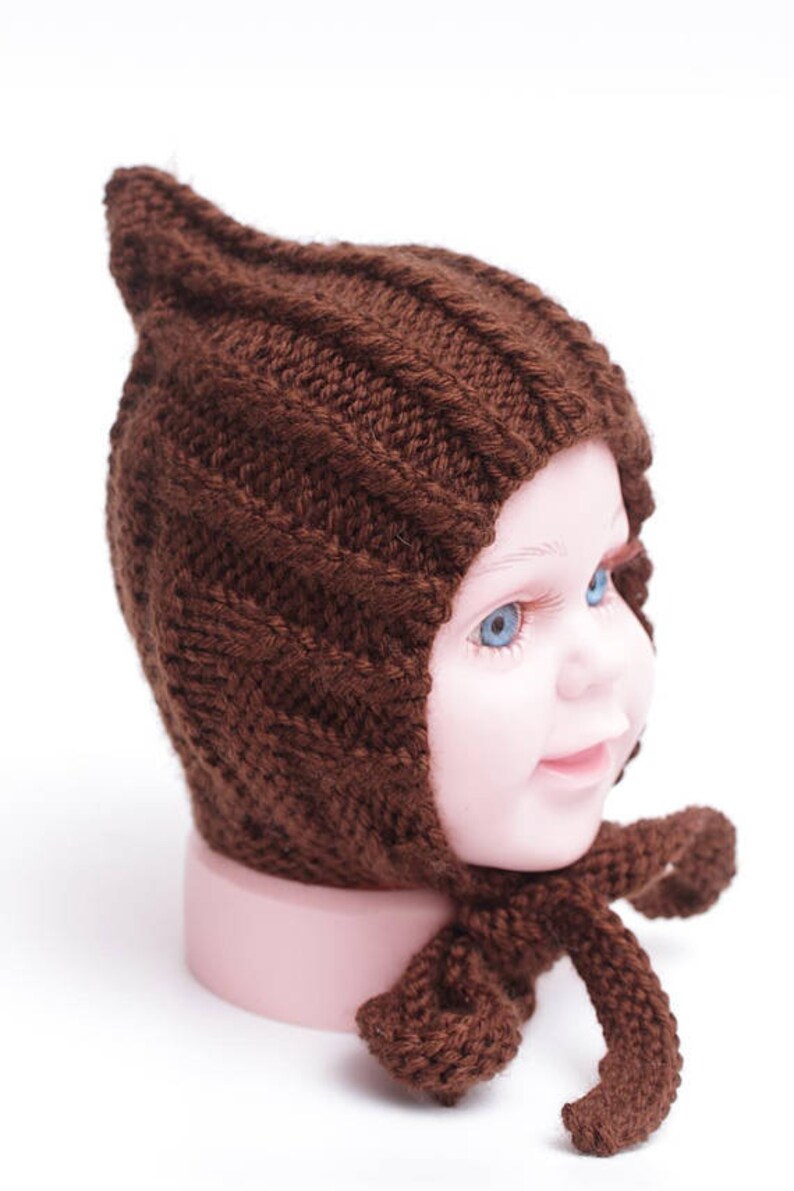 K&ouml;nnte beinhalten: Eine braune gestrickte Baby-M&uuml;tze mit einer spitzen Spitze und einer langen Schleife. Die M&uuml;tze sitzt auf einem Mannequin-Kopf.
