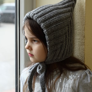 Op de afbeelding: Een jong meisje draagt een grijze gebreide capuchon met een lange, gebonden sjaal. De muts heeft een puntige top en een geribbelde textuur. Het meisje kijkt naar de zijkant.