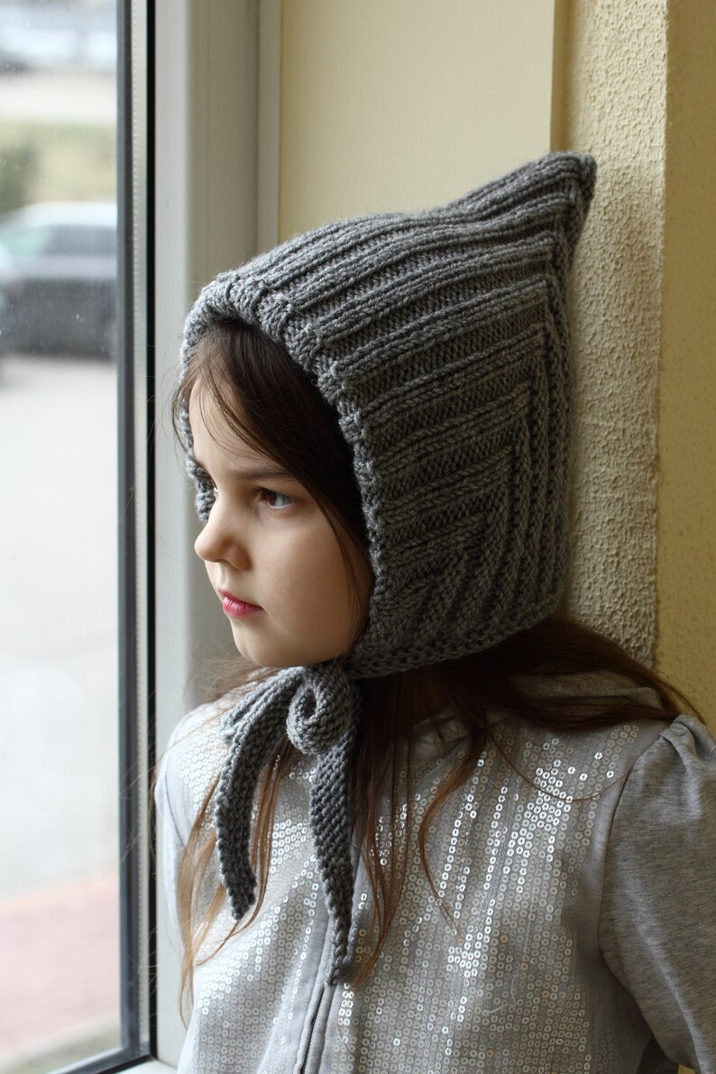 Op de afbeelding: Een jong meisje draagt een grijze gebreide capuchon met een lange, gebonden sjaal. De muts heeft een puntige top en een geribbelde textuur. Het meisje kijkt naar de zijkant.