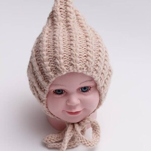 Peut inclure: Un bonnet b&eacute;b&eacute; en tricot beige avec un sommet pointu et une longue cravate. Le bonnet est fait d'un fil doux et &eacute;pais et a un design simple et classique.