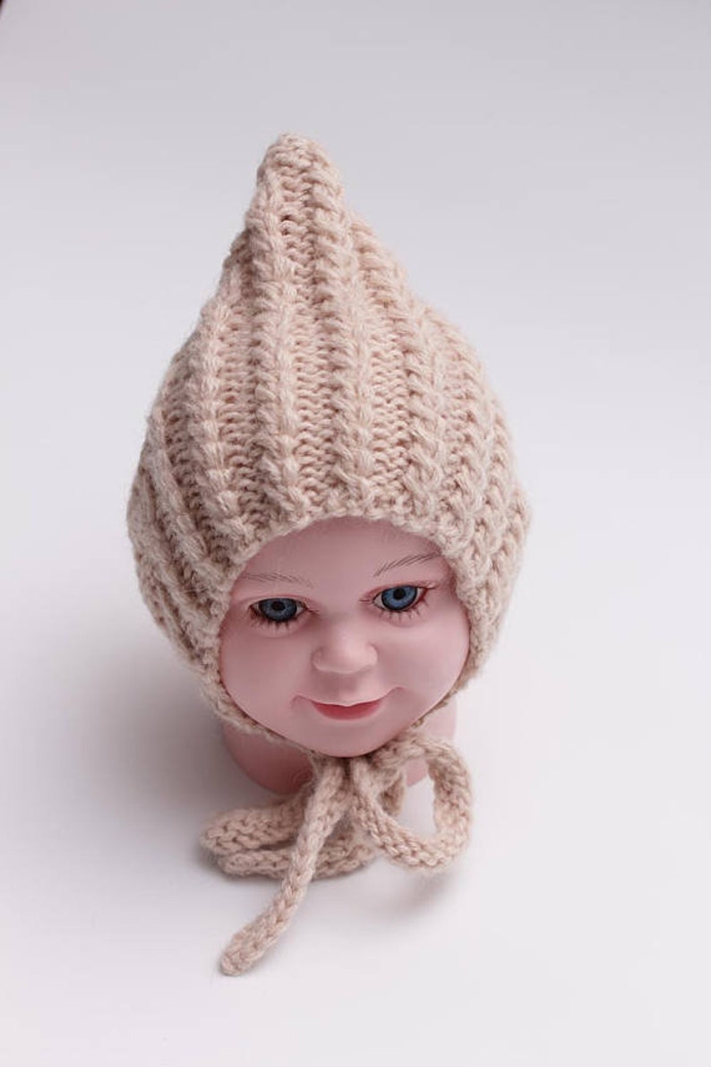 Peut inclure: Un bonnet b&eacute;b&eacute; en tricot beige avec un sommet pointu et une longue cravate. Le bonnet est fait d'un fil doux et &eacute;pais et a un design simple et classique.