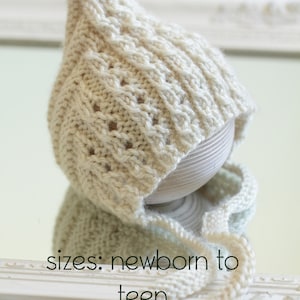 May include: A cream knitted hat with a cable stitch pattern. The hat is displayed on a wooden stand and has a long, thin tie. The text "sizes: newborn to teen" is visible below the hat.