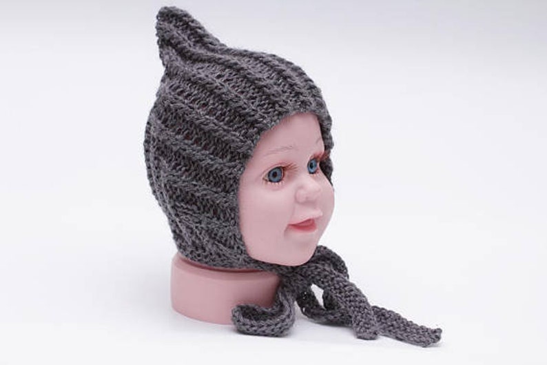May include: A gray knitted baby bonnet with a pointed top and a long tie. The bonnet is made of a soft, textured yarn and has a simple, classic design.