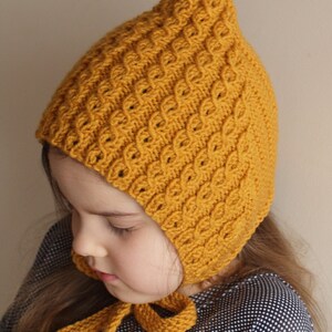 May include: A child wearing a yellow knitted hat with a pointed top and a long tie. The hat has a textured pattern and is tied under the chin.