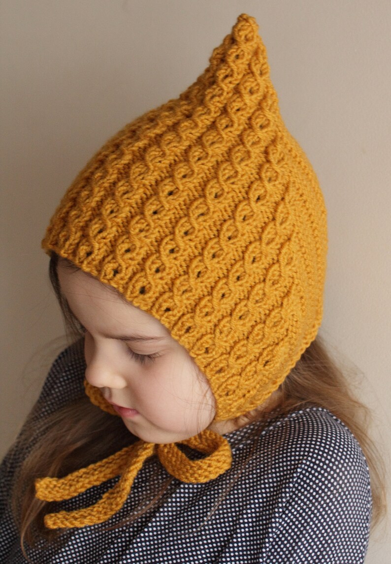 May include: A child wearing a yellow knitted hat with a pointed top and a long tie. The hat has a textured pattern and is tied under the chin.