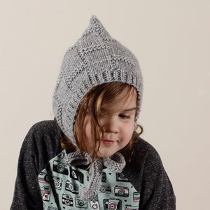 Peut inclure: Un bonnet gris en tricot avec un sommet pointu, port&eacute; par un enfant aux cheveux bruns. Le bonnet a un design unique avec un motif textur&eacute;. L'enfant porte une chemise avec un motif color&eacute; d'appareils photo.
