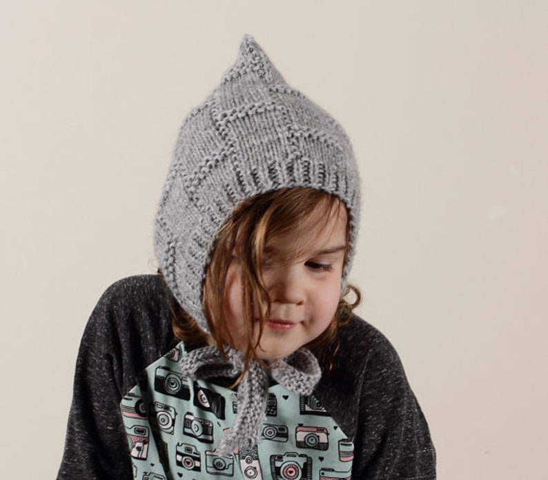 Peut inclure: Un bonnet gris en tricot avec un sommet pointu, port&eacute; par un enfant aux cheveux bruns. Le bonnet a un design unique avec un motif textur&eacute;. L'enfant porte une chemise avec un motif color&eacute; d'appareils photo.