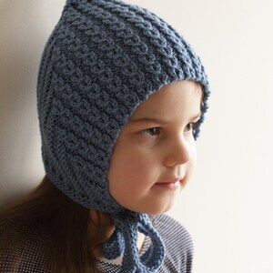 Pu&ograve; includere: Un bambino che indossa un cappello di lana blu con la cima a punta e un lungo laccio. Il cappello ha un motivo testurizzato ed &egrave; legato sotto il mento.