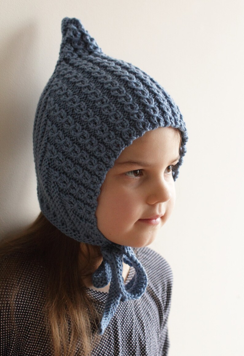 Pu&ograve; includere: Un bambino che indossa un cappello di lana blu con la cima a punta e un lungo laccio. Il cappello ha un motivo testurizzato ed &egrave; legato sotto il mento.
