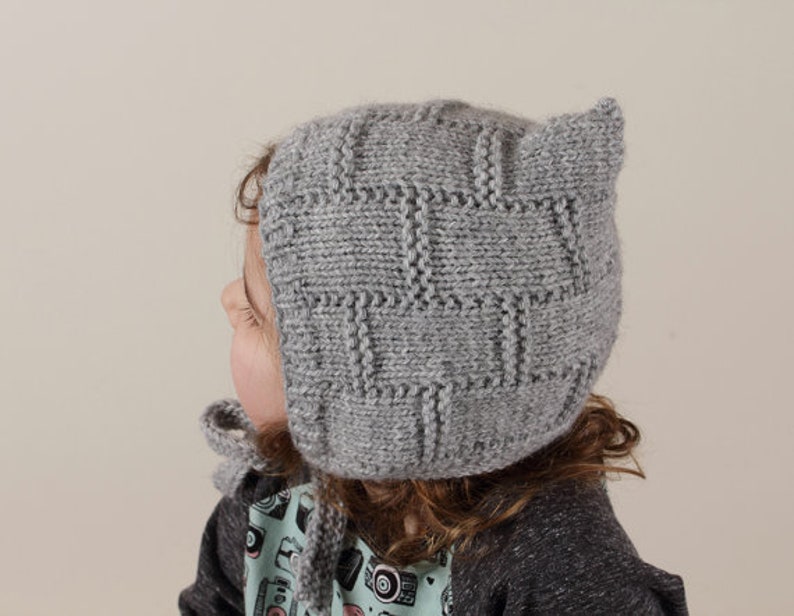 Peut inclure: Un bonnet b&eacute;b&eacute; en tricot gris avec des rabats d'oreilles. Le bonnet a un motif textur&eacute; et est fait de fil doux.