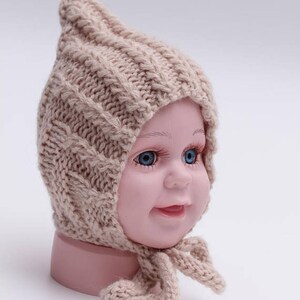 Peut inclure: Un bonnet b&eacute;b&eacute; en tricot beige avec un sommet pointu et une longue cravate. Le bonnet est fait d'un fil doux et &eacute;pais et pr&eacute;sente un motif en tricot torsad&eacute;.