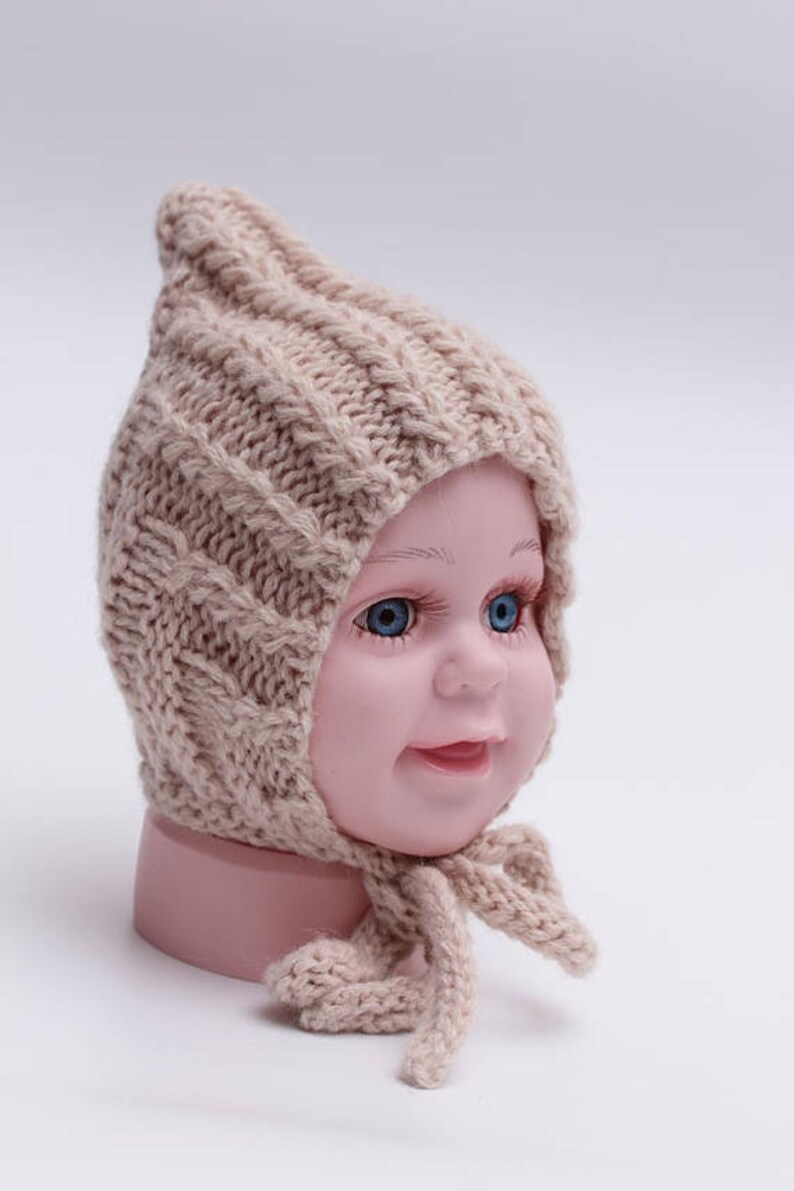 Peut inclure: Un bonnet b&eacute;b&eacute; en tricot beige avec un sommet pointu et une longue cravate. Le bonnet est fait d'un fil doux et &eacute;pais et pr&eacute;sente un motif en tricot torsad&eacute;.