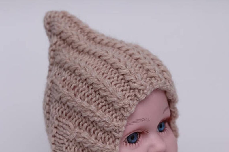 Peut inclure: Un bonnet b&eacute;b&eacute; en tricot beige avec un sommet pointu et un motif en tricot torsad&eacute;.