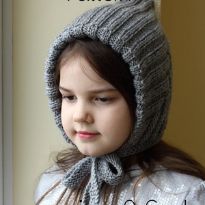 Op de afbeelding: Een grijze gebreide muts met een strik, met een geribbelde textuur. De muts wordt gedragen door een kind en de tekst "sizes: 0-6m to adult" is zichtbaar onder de muts.