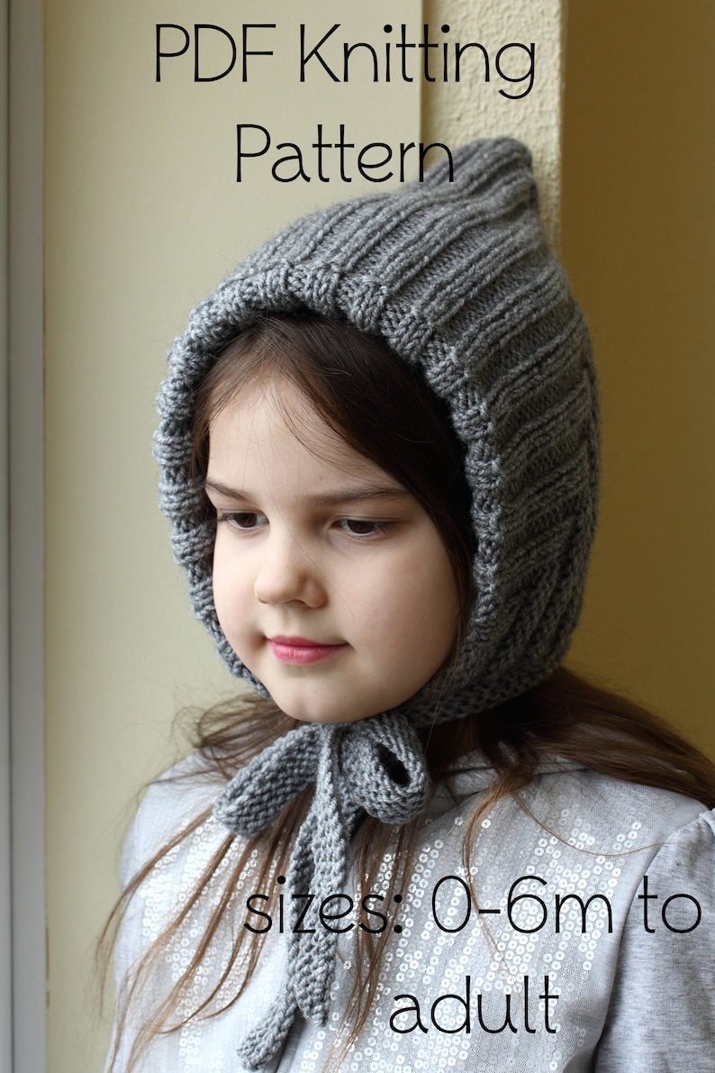 Op de afbeelding: Een grijze gebreide muts met een strik, met een geribbelde textuur. De muts wordt gedragen door een kind en de tekst "sizes: 0-6m to adult" is zichtbaar onder de muts.