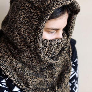 May include: A brown and black knitted hooded cowl with a drawstring. The cowl is being worn by a person, but only the cowl is visible in the image.