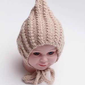 Peut inclure: Un bonnet b&eacute;b&eacute; en tricot beige avec un sommet pointu et une longue cravate. Le bonnet est fait d'un fil doux et textur&eacute; et a un design simple et classique.