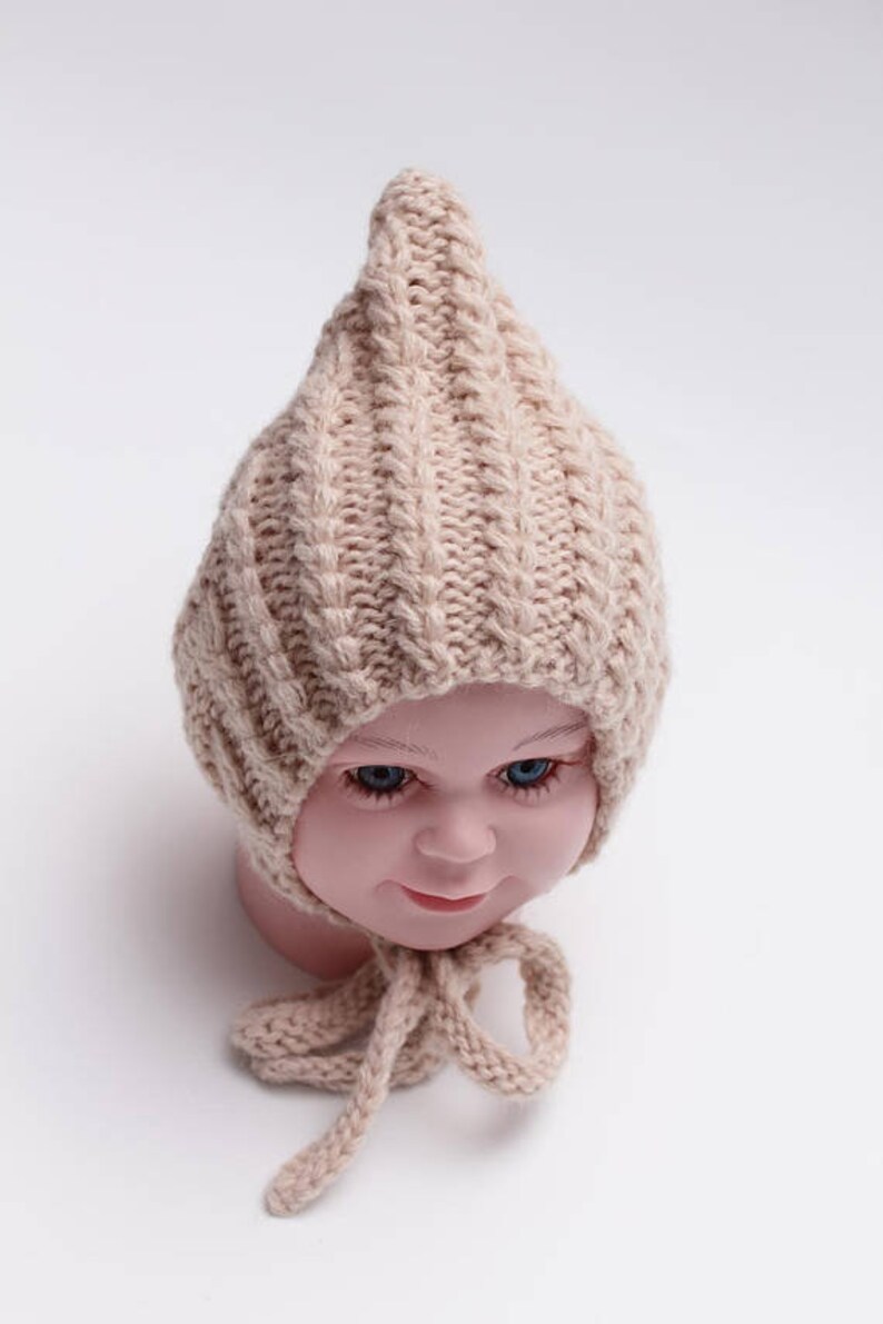 Peut inclure: Un bonnet b&eacute;b&eacute; en tricot beige avec un sommet pointu et une longue cravate. Le bonnet est fait d'un fil doux et textur&eacute; et a un design simple et classique.