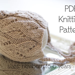 Pode incluir: Um gorro de bebé de malha em cor castanho claro com um padrão de tricô de cabo. O gorro tem um laço longo que pode ser amarrado sob o queixo. O texto "PDF Knitting Pattern" e "sizes: newborn to 6 years" é visível na imagem.