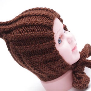 K&ouml;nnte beinhalten: Eine braune gestrickte Baby-M&uuml;tze mit einer spitzen Spitze und einer passenden Schleife. Die M&uuml;tze ist aus weichem Garn gefertigt und hat ein schlichtes, klassisches Design.