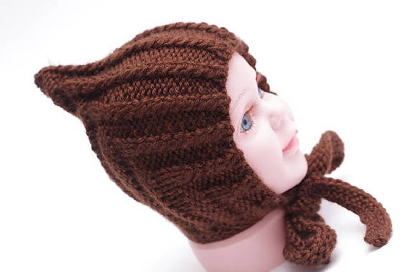 K&ouml;nnte beinhalten: Eine braune gestrickte Baby-M&uuml;tze mit einer spitzen Spitze und einer passenden Schleife. Die M&uuml;tze ist aus weichem Garn gefertigt und hat ein schlichtes, klassisches Design.