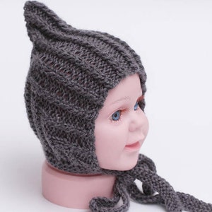 May include: A gray knitted baby hat with a pointed top and a long tie. The hat is on a mannequin head.