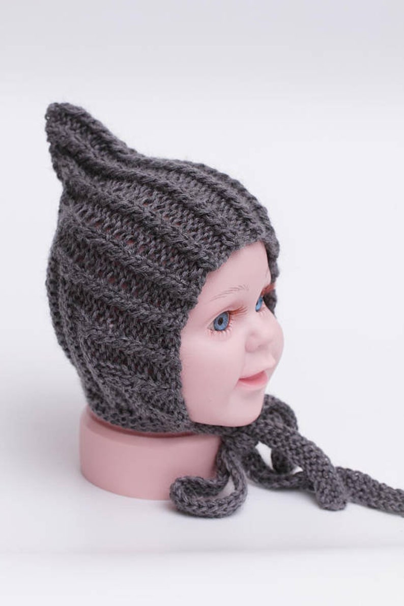 May include: A gray knitted baby hat with a pointed top and a long tie. The hat is on a mannequin head.
