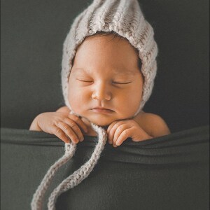 Peut inclure: Un nouveau-n&eacute; endormi portant un bonnet en tricot marron clair avec une longue cravate. Le b&eacute;b&eacute; est allong&eacute; sur un tissu gris fonc&eacute;.