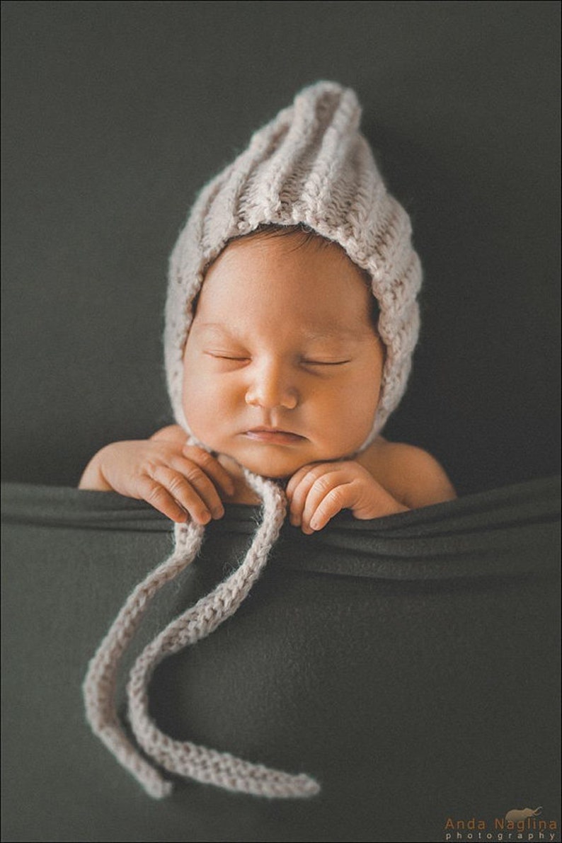 Peut inclure: Un nouveau-n&eacute; endormi portant un bonnet en tricot marron clair avec une longue cravate. Le b&eacute;b&eacute; est allong&eacute; sur un tissu gris fonc&eacute;.