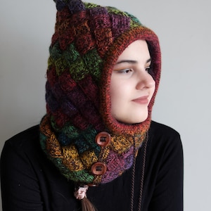 Puede incluir: Un cuello de punto multicolor con capucha a juego. El cuello tiene dos botones de madera y un cordón largo. La capucha está subida sobre la cabeza de la persona que la lleva.  Patrón de tejido PDF.