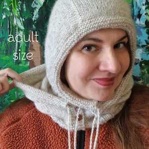 以下が含まれることがあります： ドローコードと木製ボタンが付いた、明るい灰色のニットフード。「PDF Knitting Pattern」と「adult size」の文字が見えます。フードは錆色のジャケットの上に着用されています。