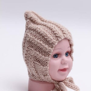 Peut inclure: Un bonnet b&eacute;b&eacute; en tricot beige avec un sommet pointu et une longue cravate. Le bonnet est fait d'un fil doux et &eacute;pais et pr&eacute;sente un motif en tricot torsad&eacute;.
