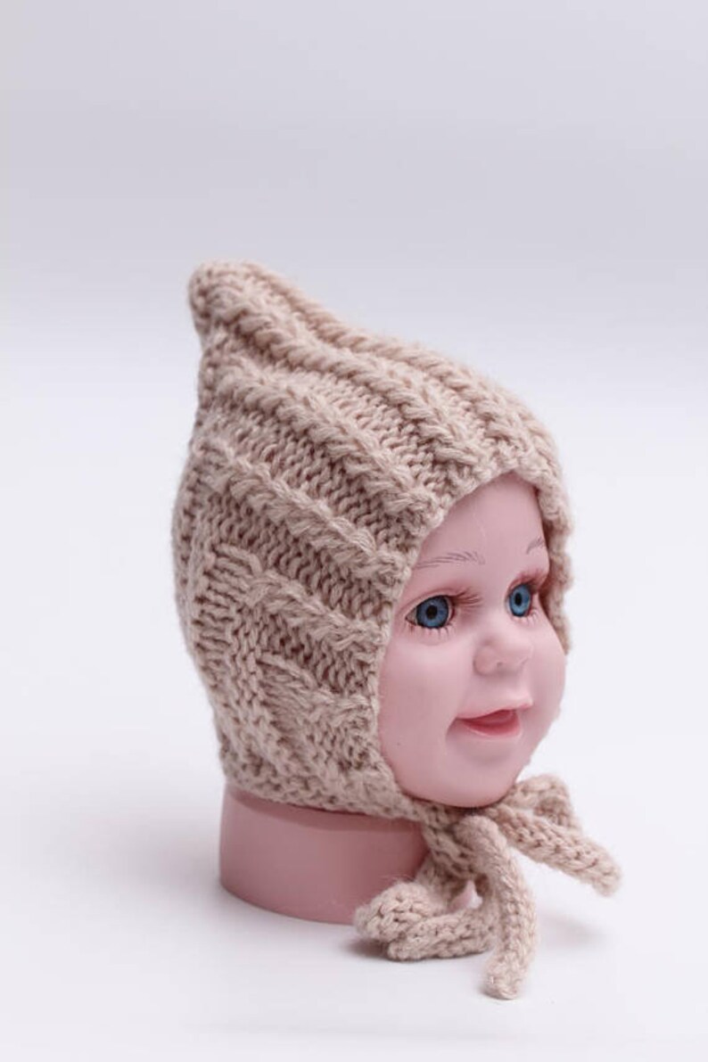 Peut inclure: Un bonnet b&eacute;b&eacute; en tricot beige avec un sommet pointu et une longue cravate. Le bonnet est fait d'un fil doux et &eacute;pais et pr&eacute;sente un motif en tricot torsad&eacute;.