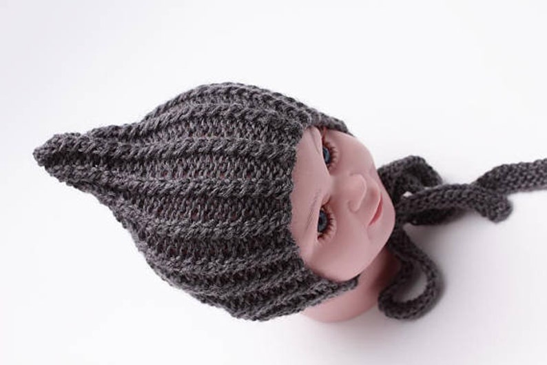 May include: Gray knitted baby hat with a pointed top and a matching scarf. The hat has a ribbed texture and is tied with a long scarf.