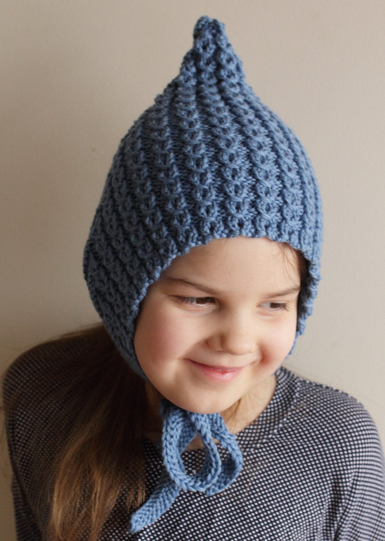 Pu&ograve; includere: Un cappello blu a maglia con la cima a punta e le alette per le orecchie. Il cappello ha un design semplice e testurizzato e si lega sotto il mento.