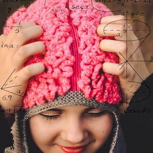 Puede incluir: Una persona joven con un gorro de punto rosa y gris con las manos en la cabeza. El fondo es una pizarra con ecuaciones y fórmulas matemáticas escritas con tiza blanca.
