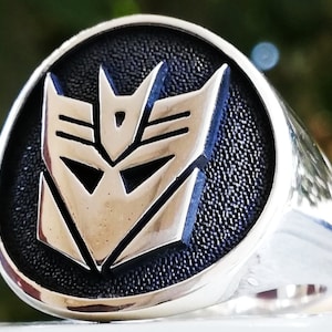 Puede incluir: Un anillo de plata con fondo negro que presenta el logotipo de los Decepticons de Transformers. El logotipo es una cara de robot de plata estilizada con un diseño angular y afilado.