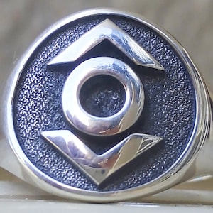 May include: A silver ring with a circular face featuring a central circle, an upward-pointing chevron, and a downward-pointing chevron. The ring's face has a textured, dark background, and the design elements are polished silver.