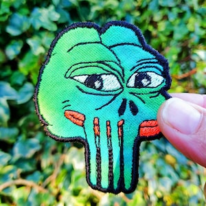 May include: Green and black embroidered patch of a skull with a green frog face, red lips, and black eyes. The skull has a green and black striped pattern.