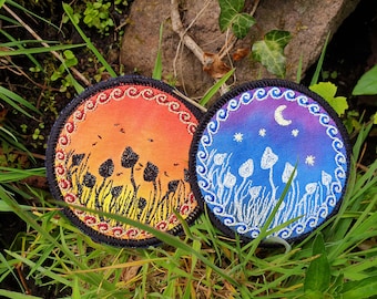 May include: Two round embroidered patches with a mushroom theme. One patch features a sunset with orange, red, and yellow colors and black mushrooms. The other patch features a night sky with blue, purple, and white colors and white mushrooms.