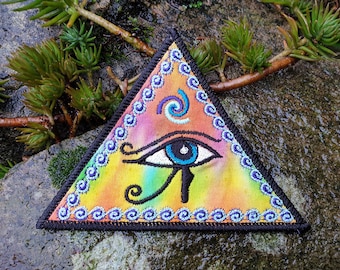 May include: A triangular patch with a colorful tie-dye background and an embroidered eye with a blue iris and black pupil. The eye is surrounded by a swirling design and a border of blue and purple swirls.