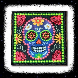 May include: Square patch featuring a colorful sugar skull design. The skull is blue with embroidered details, including flowers, roses, and a smiling mouth. The patch has a black border with green stitching and is suitable for clothing or accessories.