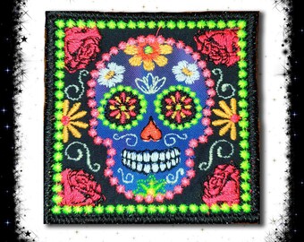May include: Square patch featuring a colorful sugar skull design. The skull is blue with embroidered details, including flowers, roses, and a smiling mouth. The patch has a black border with green stitching and is suitable for clothing or accessories.