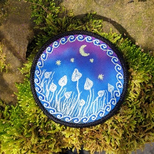 May include: A round, embroidered patch with a night sky scene. The sky is a gradient of purple and blue with white stars and a crescent moon. White mushrooms are embroidered on the ground in front of the sky.