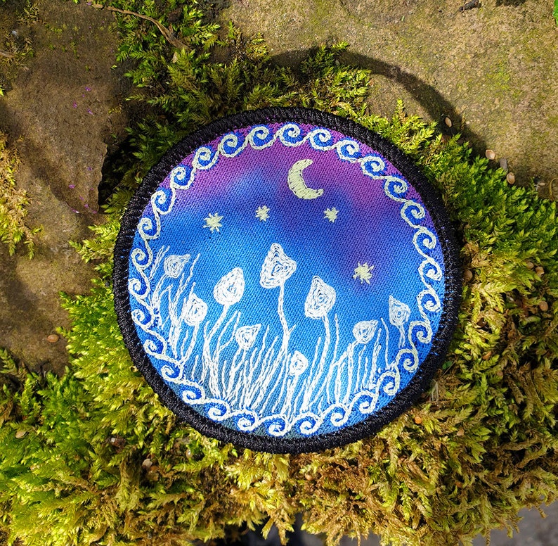May include: A round, embroidered patch with a night sky scene. The sky is a gradient of purple and blue with white stars and a crescent moon. White mushrooms are embroidered on the ground in front of the sky.